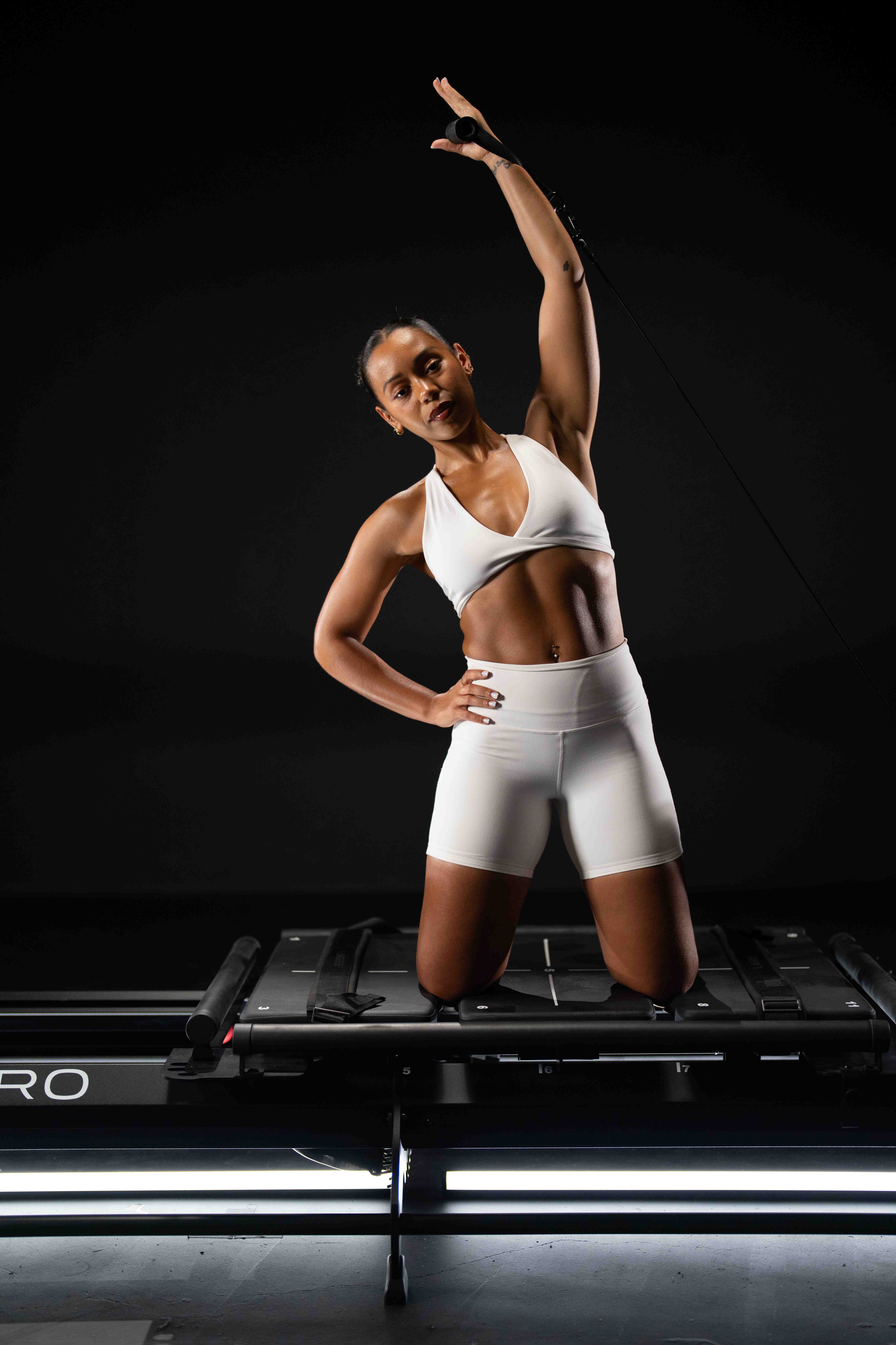 Woman in white sports bra and shorts kneeling on Mega Pro Megaformer with one arm raised pulling a resistance cable